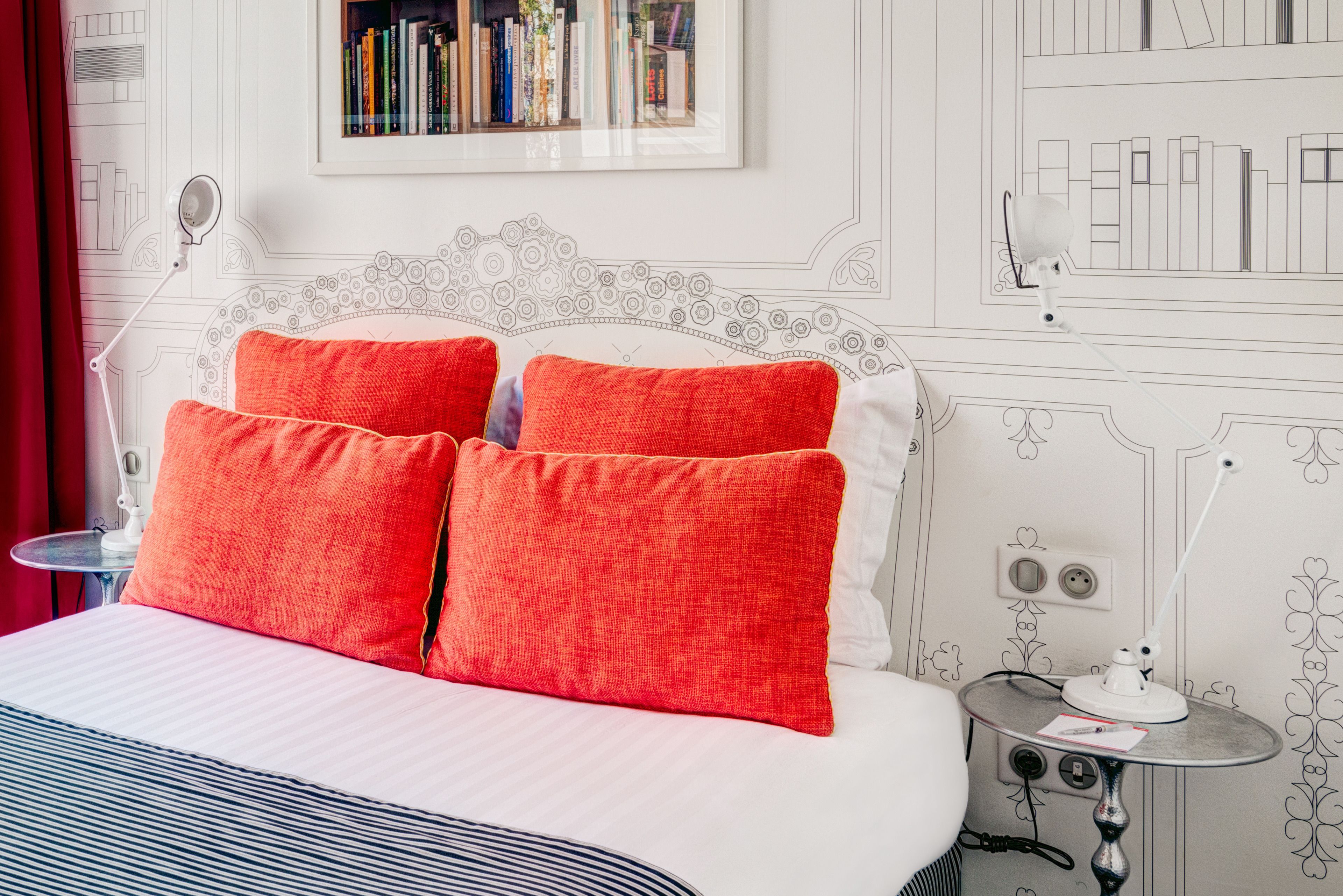A neatly made bed with red pillows, a bedside table with a lamp, and a bookshelf with books.