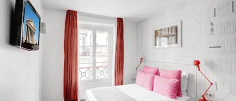 A hotel room with a bed, a TV mounted on the wall, a window with curtains, a bedside table with a lamp, and a wall with framed pictures.