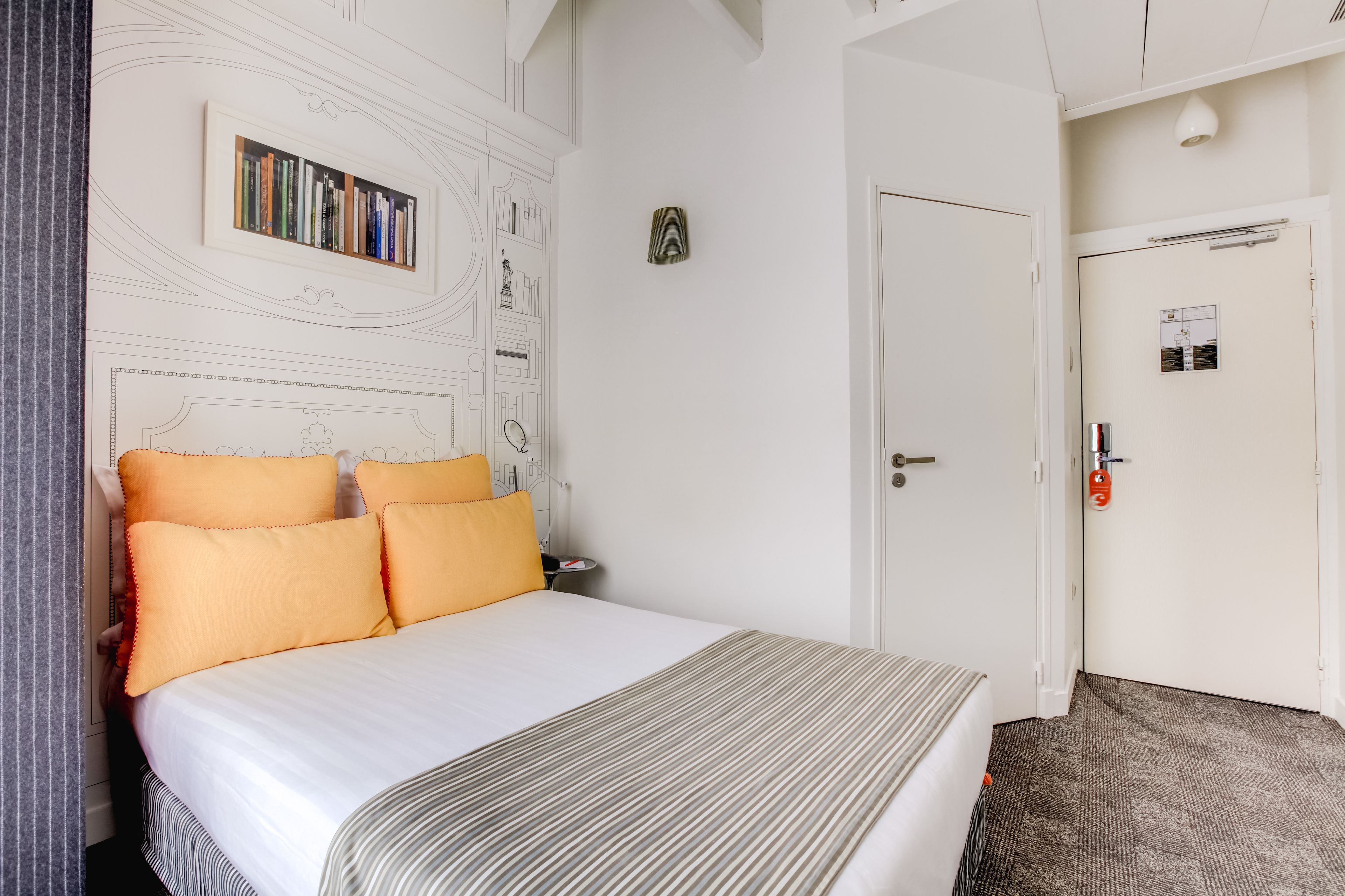Hotellihuone, jossa on suuri sänky, koristekuvioitu pääty, kirjahylly kirjoineen, palovaroittimella varustettu ovi ja matolla peitetty lattia.