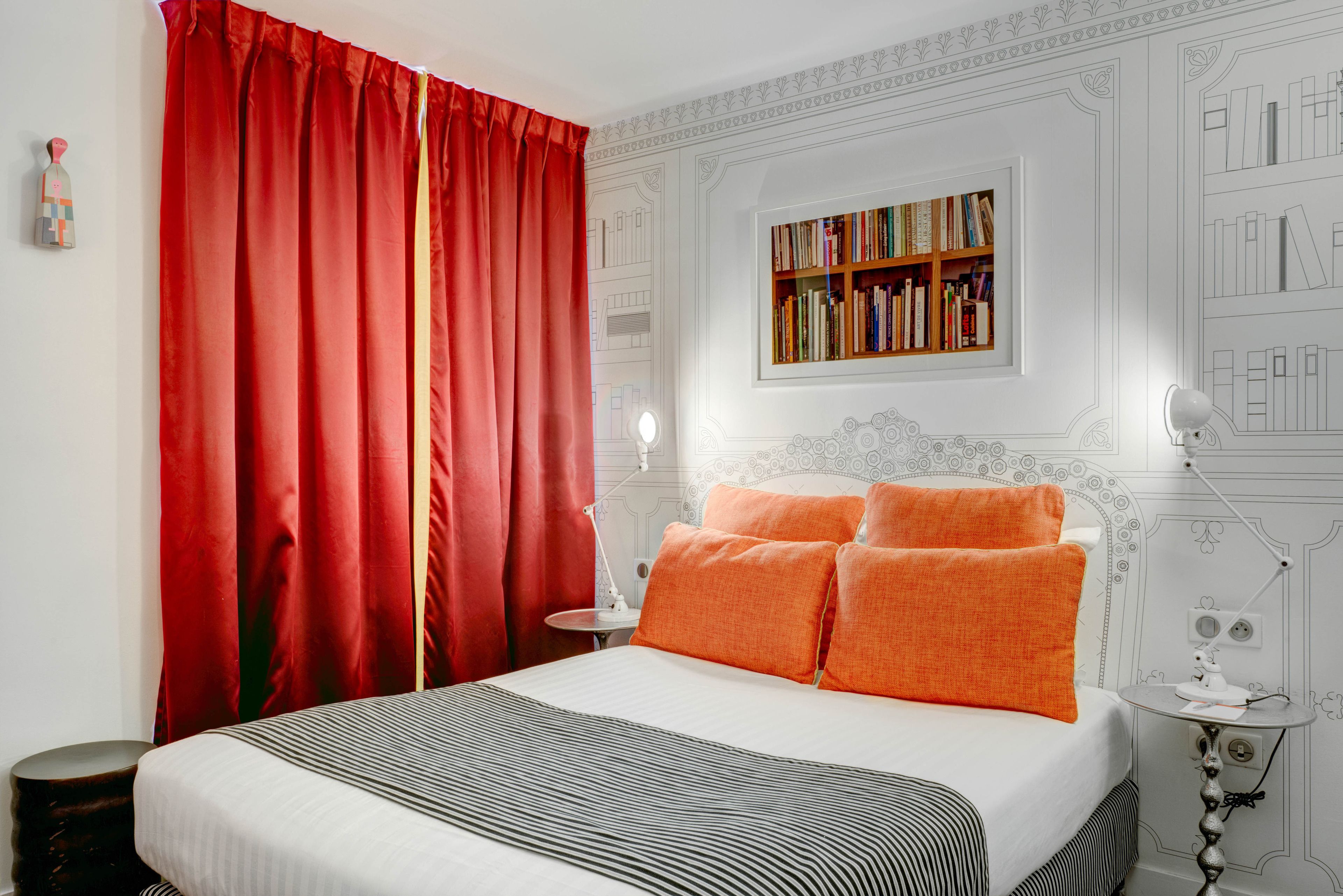 Een netjes ingerichte slaapkamer met een groot bed, rode gordijnen, een boekenkast en een klein tafeltje met een lamp.