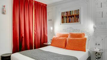 Een netjes ingerichte slaapkamer met een groot bed, rode gordijnen, een boekenkast en een klein tafeltje met een lamp.