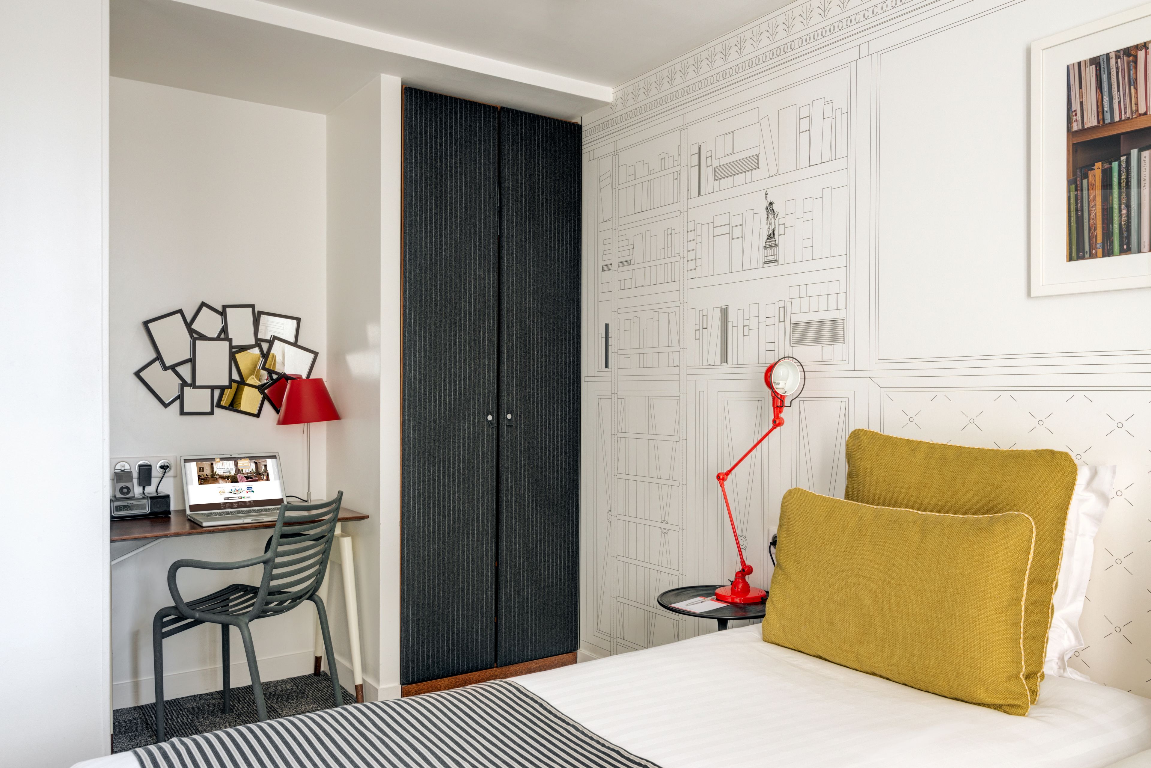 A hotel room with a bed, a desk with a laptop, a chair, a wardrobe, and a red floor lamp.