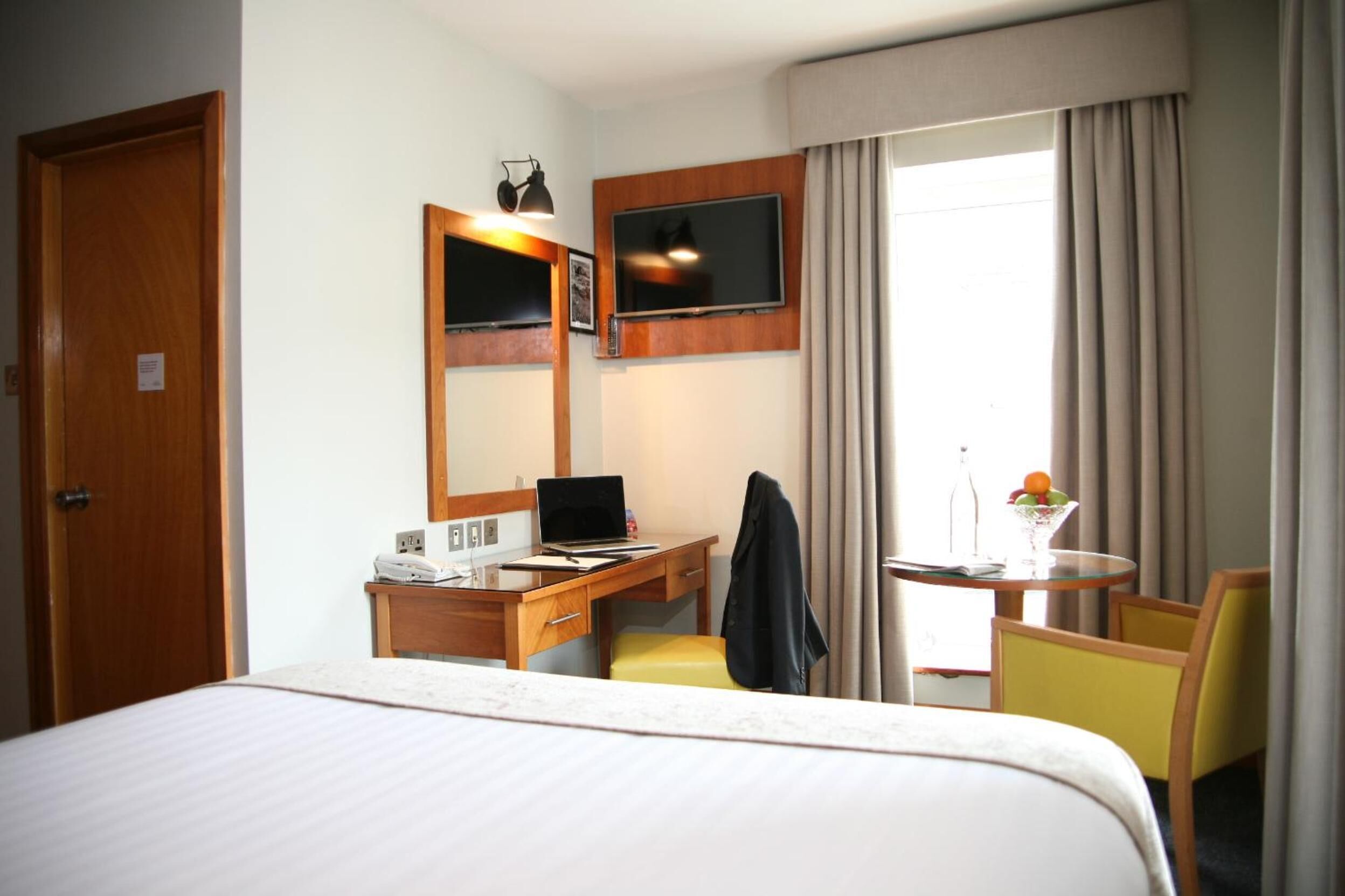 Una habitación de hotel con una cama, un escritorio con una computadora portátil, una silla, una mesita con un frutero y un televisor montado en la pared.