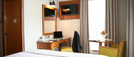 Una habitación de hotel con una cama, un escritorio con una computadora portátil, una silla, una mesita con un frutero y un televisor montado en la pared.