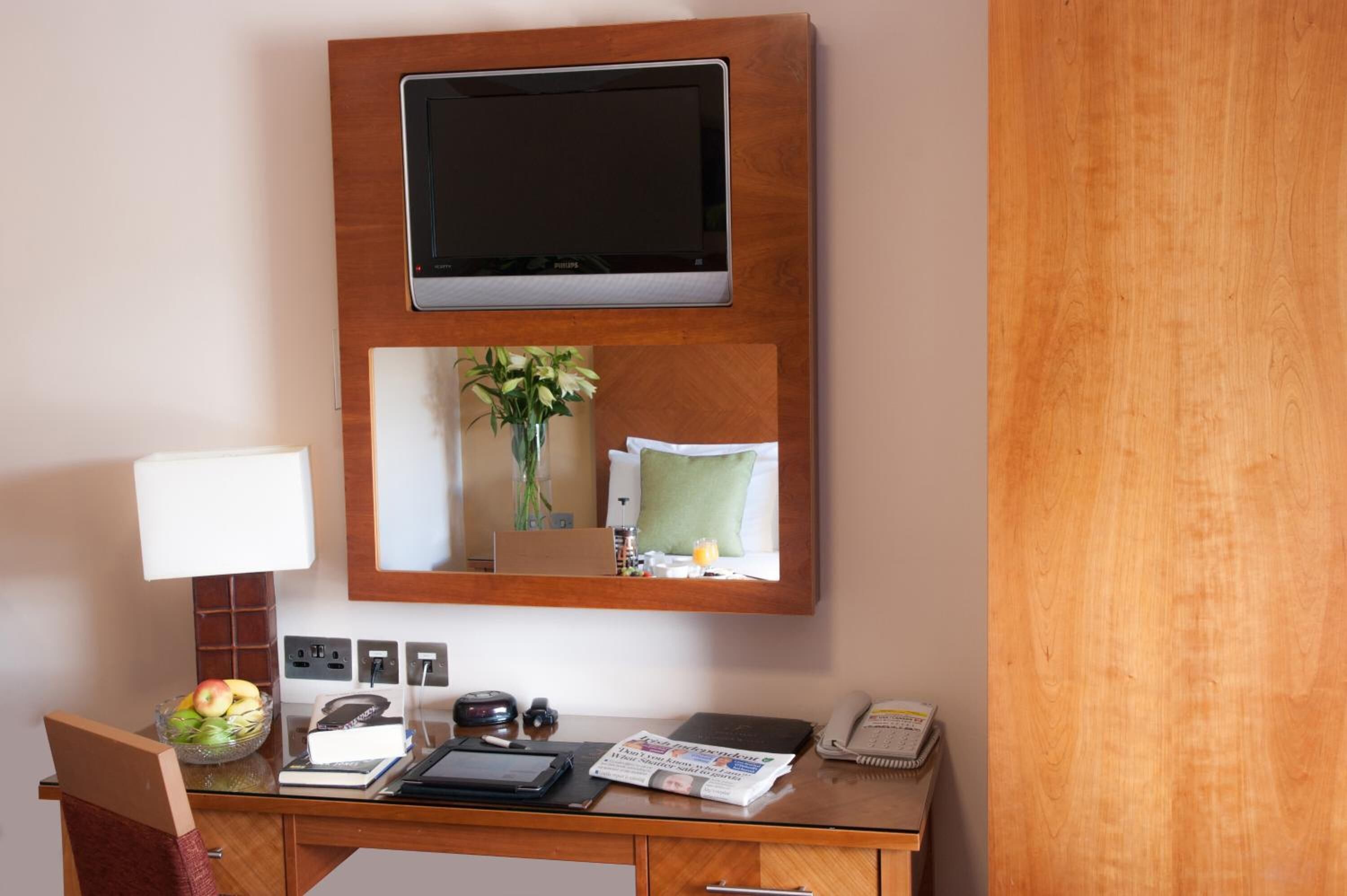 A hotel room with a wooden desk, a flat-screen TV mounted above it, a mirror reflecting the room, a vase with flowers, a lamp, a bowl of fruit, a telephone, and a magazine.