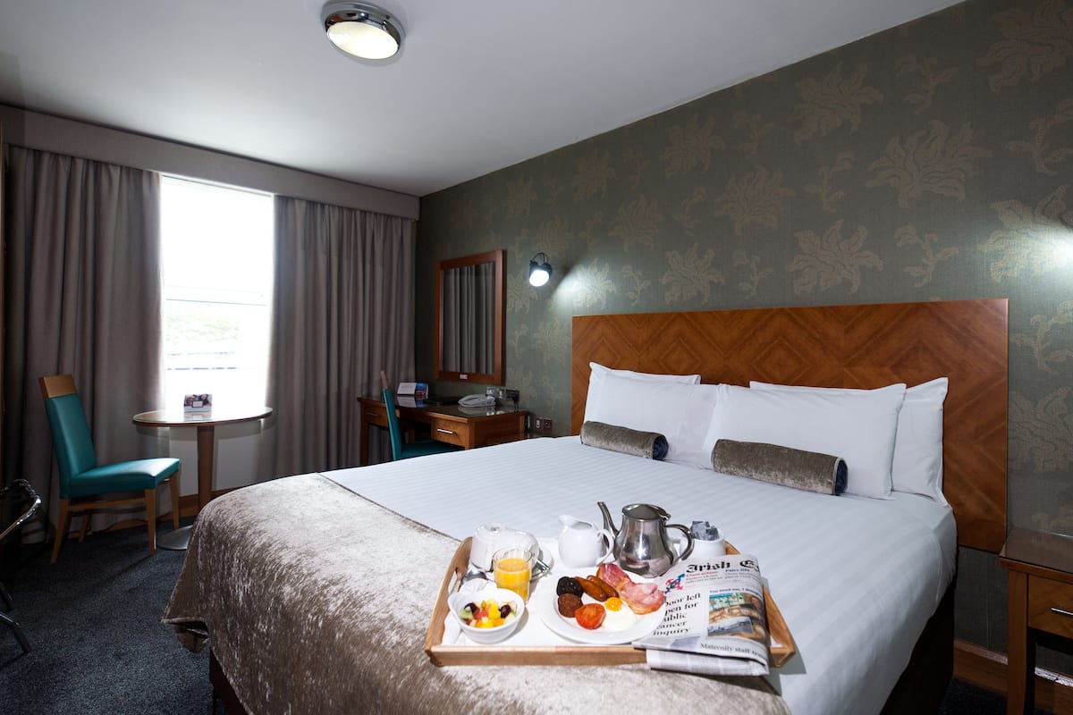Ein Hotelzimmer mit einem großen Bett, einem Nachttisch, einem Schreibtisch mit Stuhl, einem Fenster mit Vorhängen und einem Tablett mit Frühstücksangeboten.