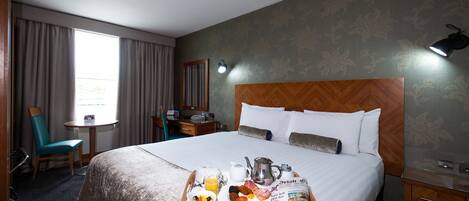 A hotel room with a large bed, a bedside table, a desk with a chair, a window with curtains, and a tray with breakfast items.