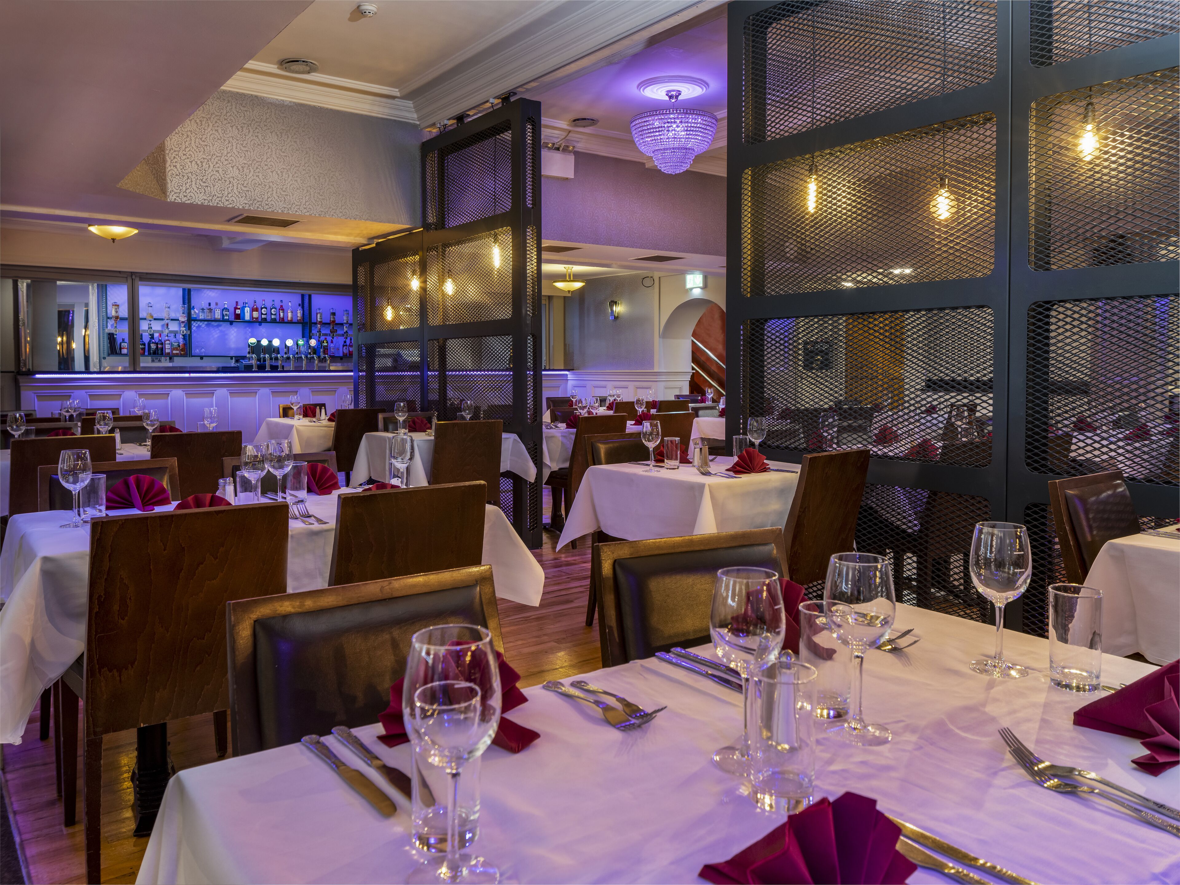 Un restaurant avec des tables dressées pour le service, un bar avec des bouteilles exposées et un luminaire décoratif au plafond.