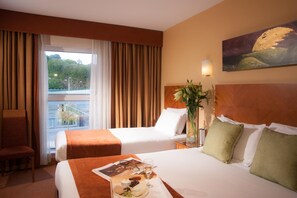 Ein Hotelzimmer mit zwei Betten, einem großen Fenster mit Vorhängen, einem Bild an der Wand und einem Nachttisch mit Blumenvase.