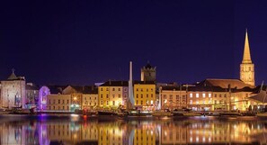 Ein beleuchtetes Stadtbild am Wasser bei Nacht, mit Gebäuden, einem markanten Turm und einem ruhigen Fluss, der die Lichter spiegelt.