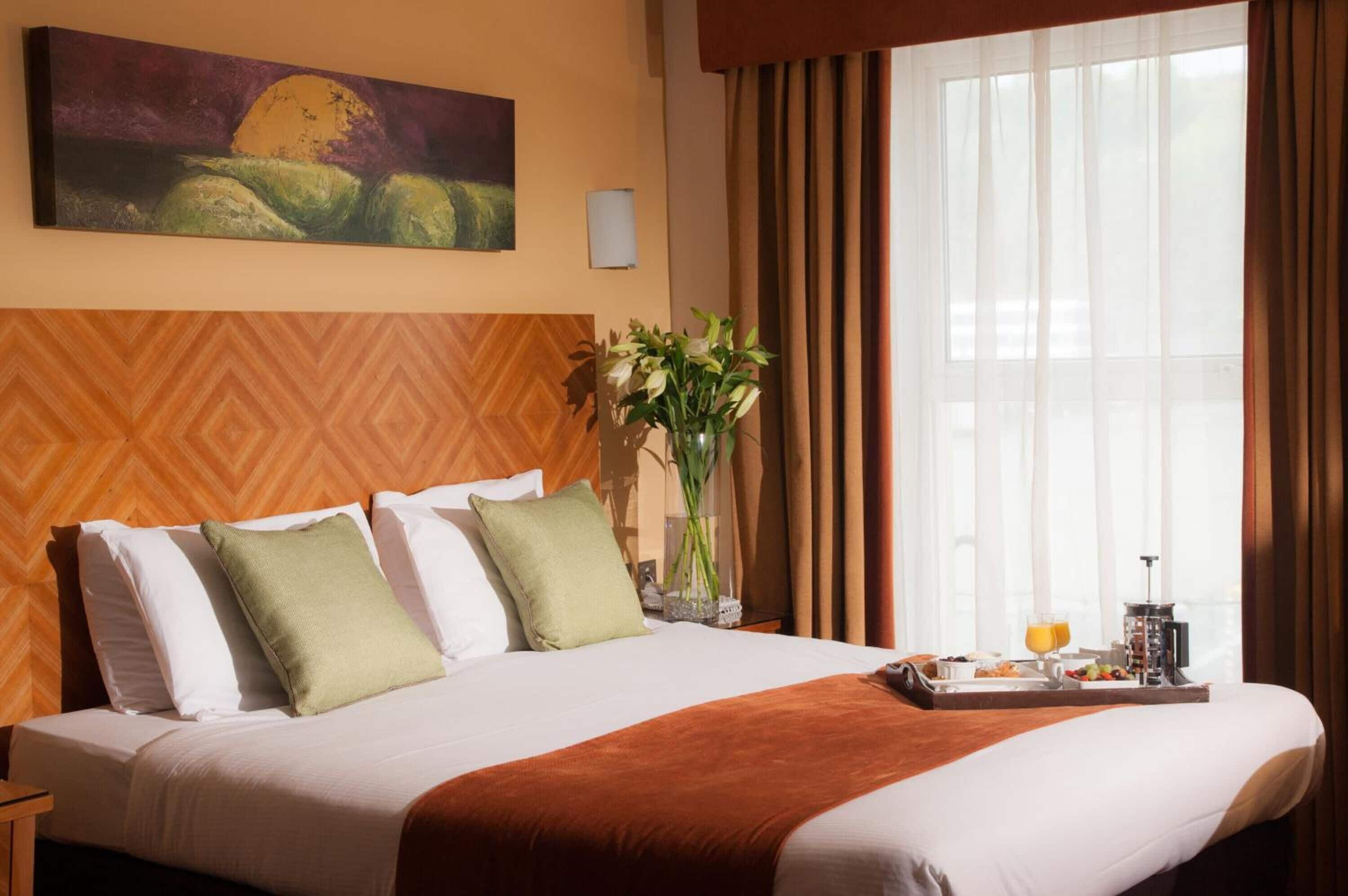 Una habitación de hotel con una cama grande, un cuadro en la pared, un florero con flores y una bandeja de desayuno en la mesita de noche.