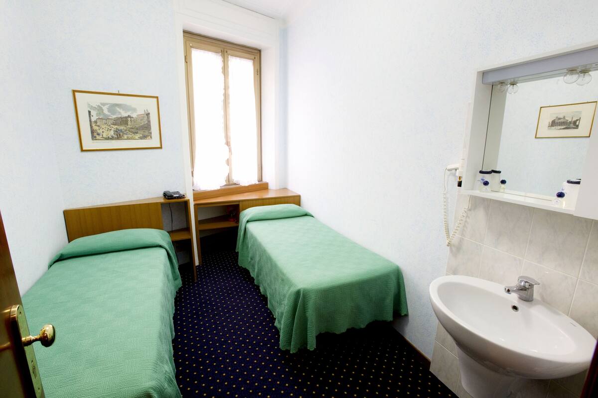 A small, clean room with two single beds, a window, a sink, and a framed picture on the wall.