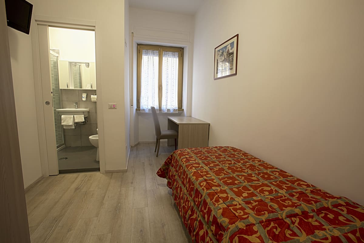 A bedroom with a bed, a desk, and a chair. There is a bathroom with a sink and toilet visible through an open door.