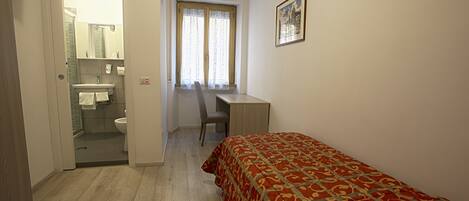 A bedroom with a bed, a desk, and a chair. There is a bathroom with a sink and toilet visible through an open door.