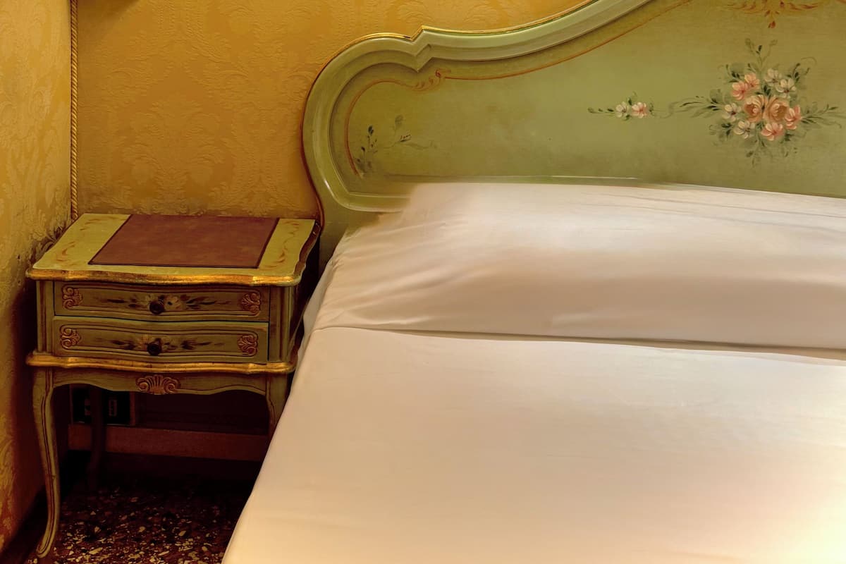 A room with a bed, a nightstand, a wall-mounted lamp, and a floral-patterned wall.