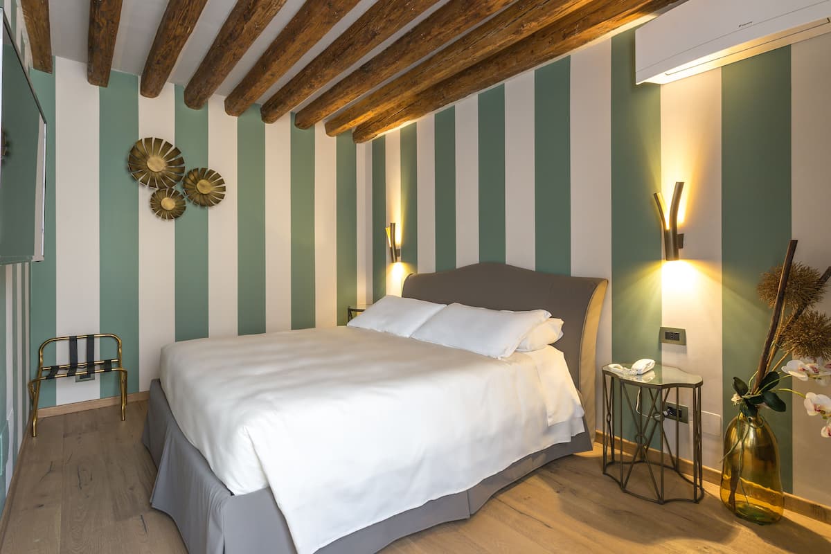 A neatly made bed in a room with striped walls, a wooden ceiling, and a bedside table with a lamp and a vase.
