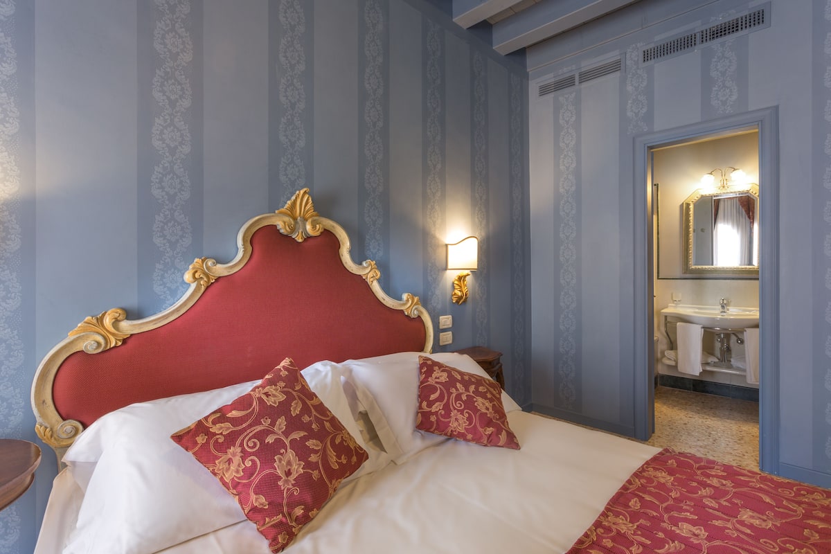 A hotel room with a red upholstered headboard, a white bedspread, and matching red and gold pillows. There is a nightstand with a lamp, and a bathroom with a sink and mirror visible through an open door.