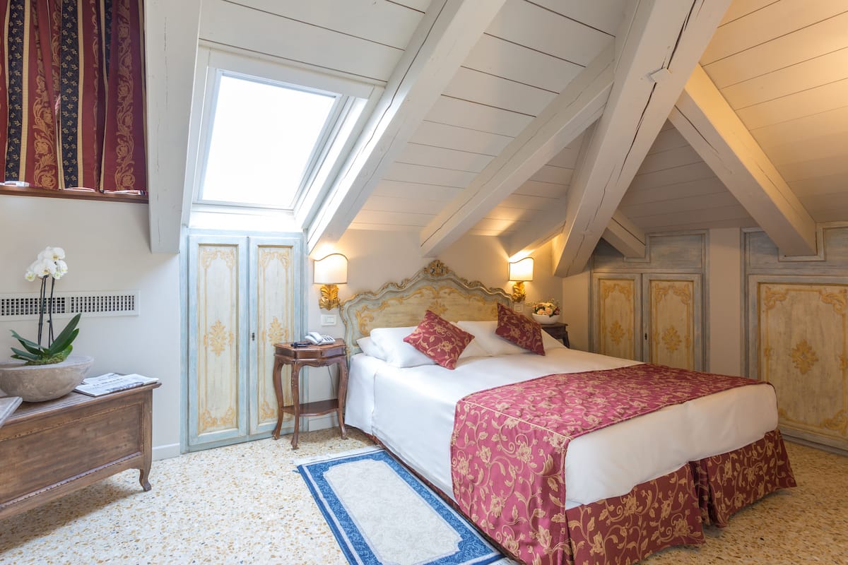 A bedroom with a large bed, a nightstand, a desk, a potted plant, and a window with curtains.