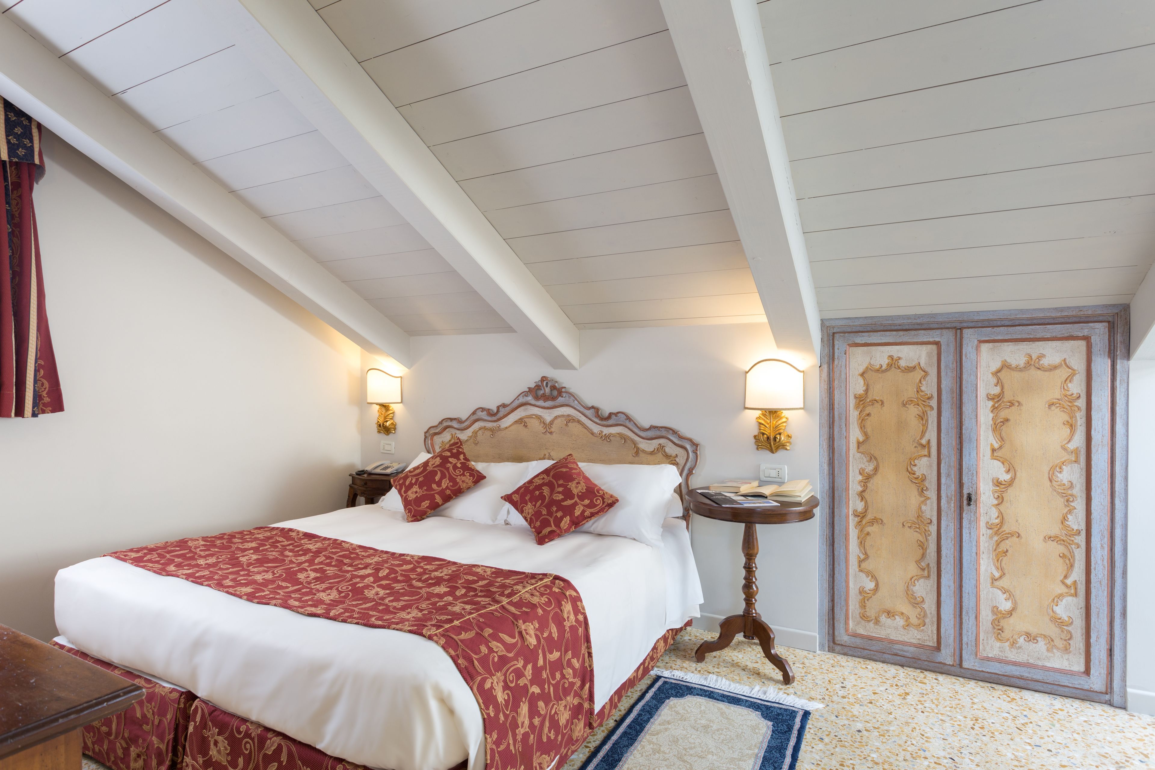 Una cama bien hecha con una colcha roja y blanca, un cabecero de madera, dos lámparas de noche y un par de puertas dobles con paneles decorativos.