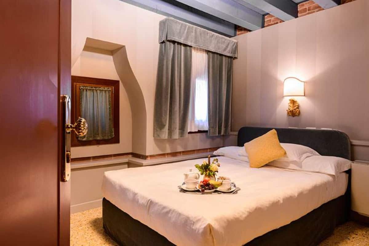 A hotel room with a bed, a bedside table, a window with curtains, and a wall-mounted light fixture.