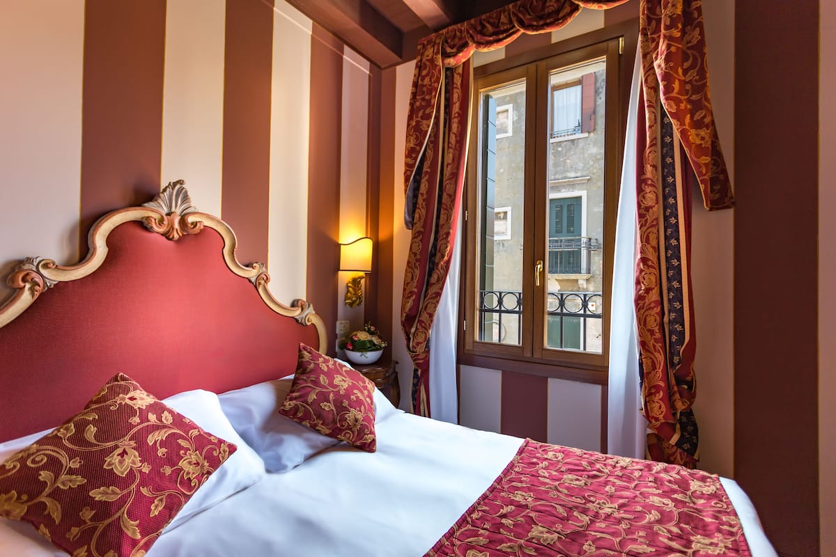 A hotel room with a large bed, red and gold patterned pillows, a white comforter, and a view of the outside balcony through a window with curtains.