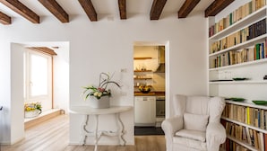 A spacious living room with a white sofa, a dining table with a vase of flowers, a bookshelf filled with books, and a kitchen in the background. - Hotel Tiziano (Venice)