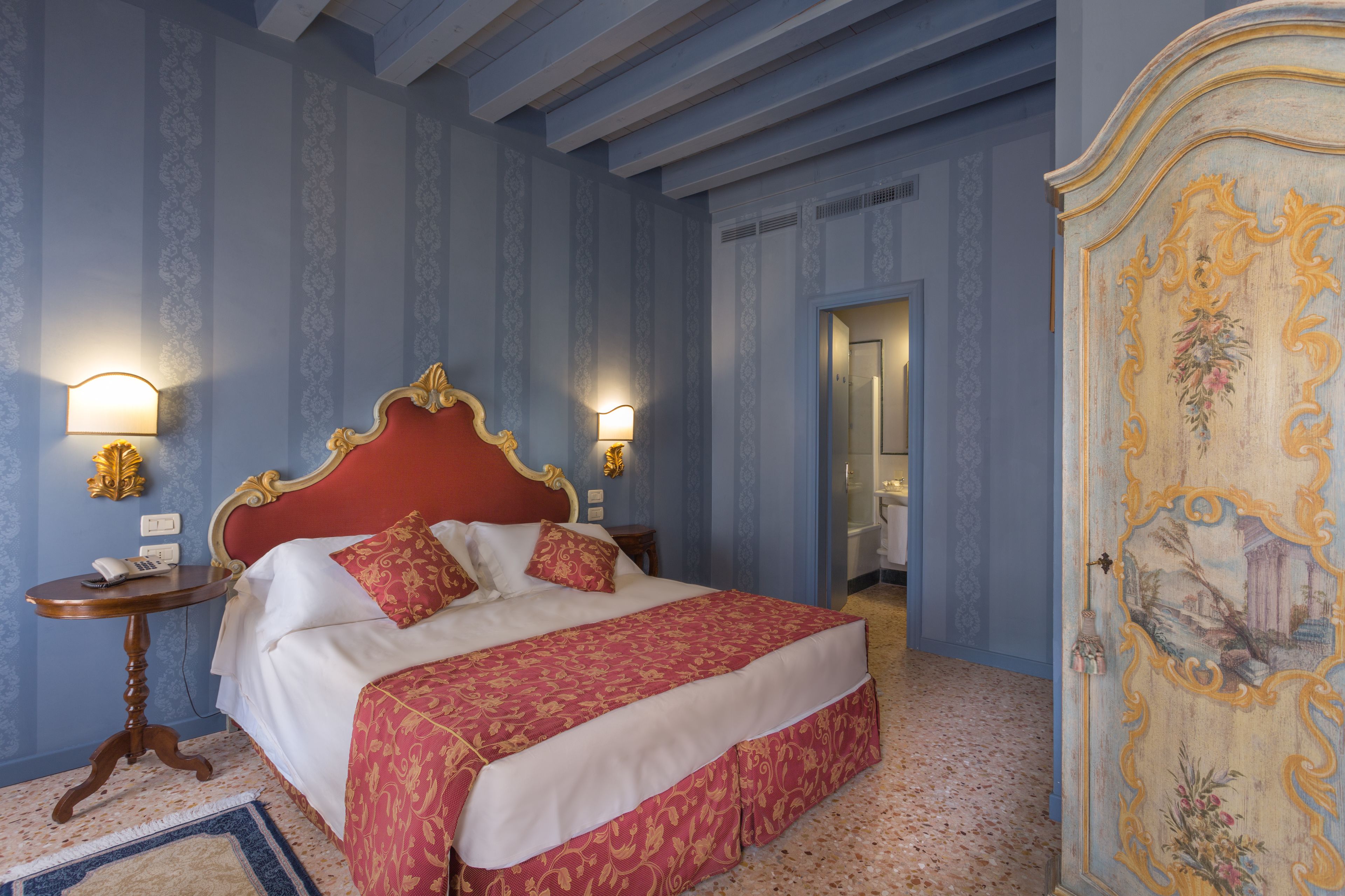 Une chambre à coucher avec une tête de lit rouge, un lit avec une literie blanche et rouge, deux tables de chevet avec des lampes, et un mur bleu orné d’un papier peint décoratif.