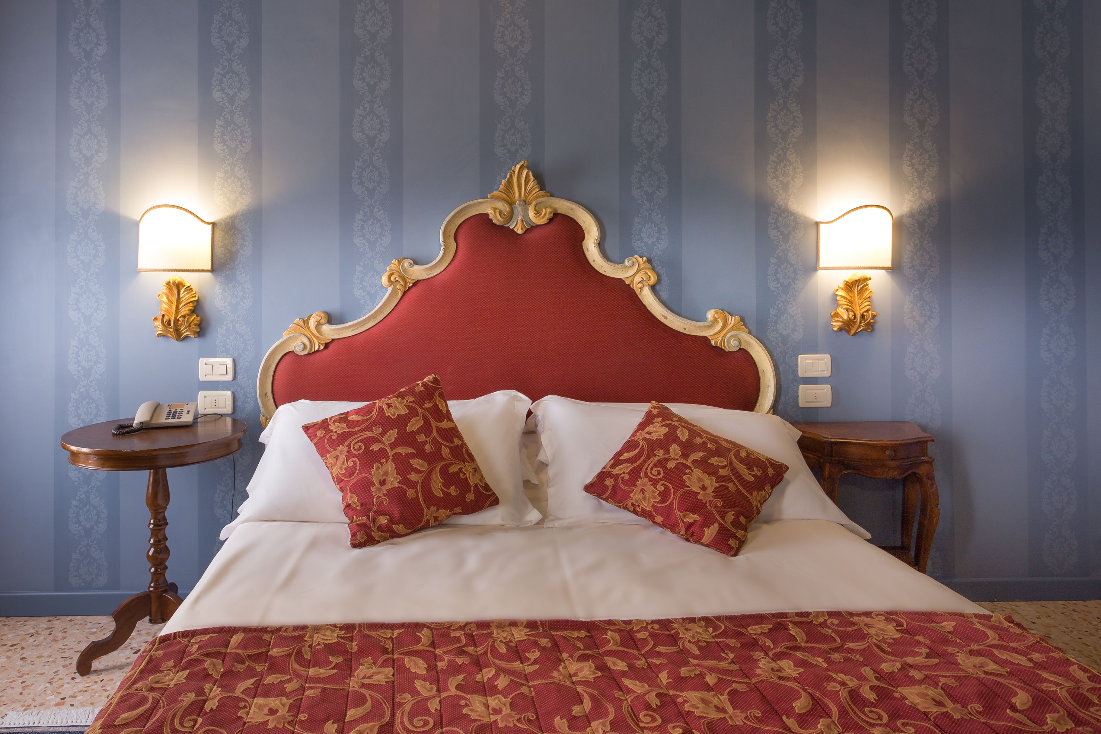 A bed with a red headboard, two matching pillows, and a patterned bedspread. There are two bedside tables with lamps and a telephone on one. The walls have a blue wallpaper with a subtle pattern.