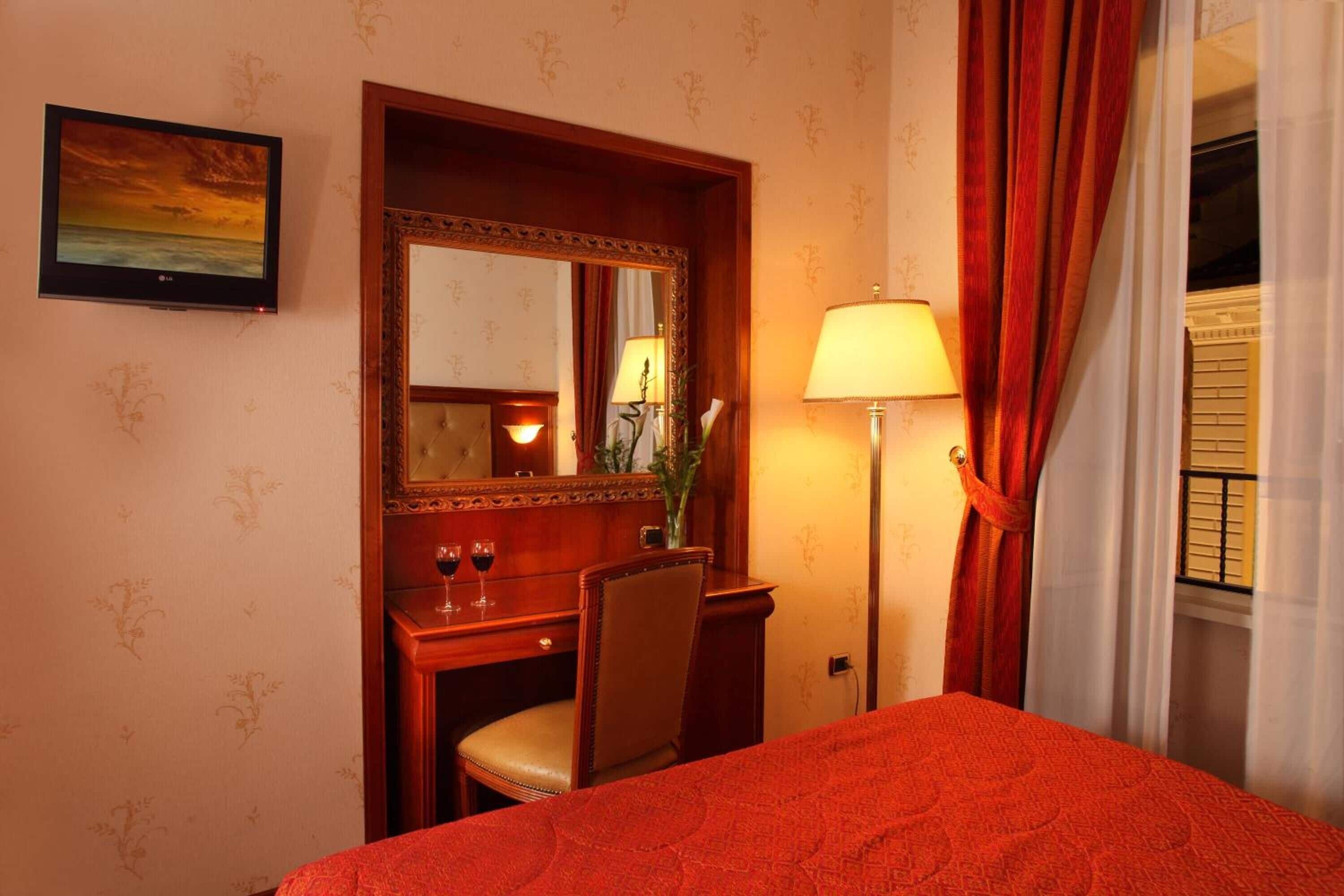 A hotel room with a bed, a desk with a chair, a mirror, a lamp, and a painting on the wall.