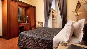 A hotel room with a large bed, a wooden wardrobe, a desk with a mirror, and a view of buildings through the window. - Serena Hotel (Rome)
