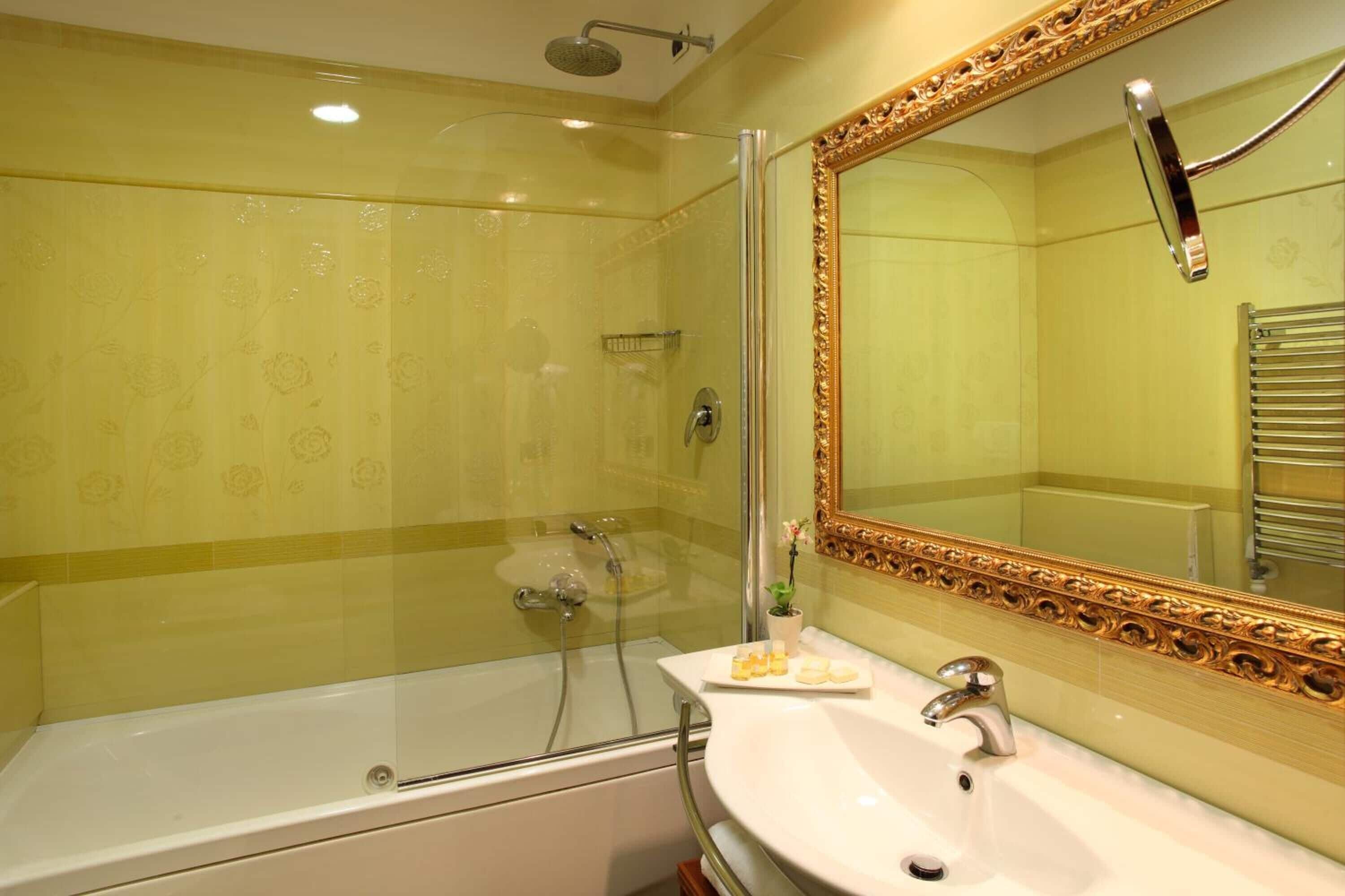 Un baño con ducha envuelta en vidrio, una bañera blanca y un tocador con espejo de marco dorado.