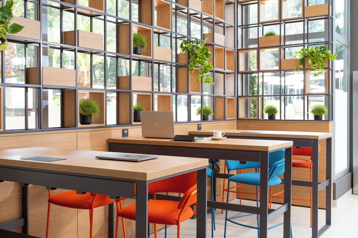 Un café moderne avec des tables en bois, des chaises orange et bleues, et une étagère où sont disposées des plantes.