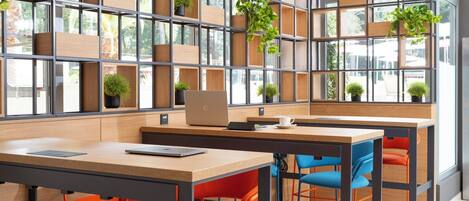 Un café moderne avec des tables en bois, des chaises orange et bleues, et une étagère où sont disposées des plantes.