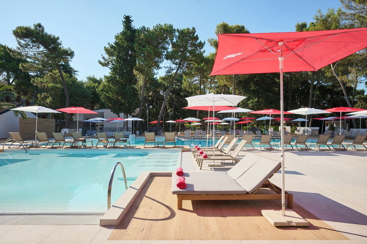 A poolside area with sun loungers and umbrellas, surrounded by trees.