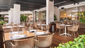 An outdoor dining area with wooden tables and chairs, stone pillars, and a view of the interior restaurant. - Hotel Pelegrin Plava Laguna (Umag)