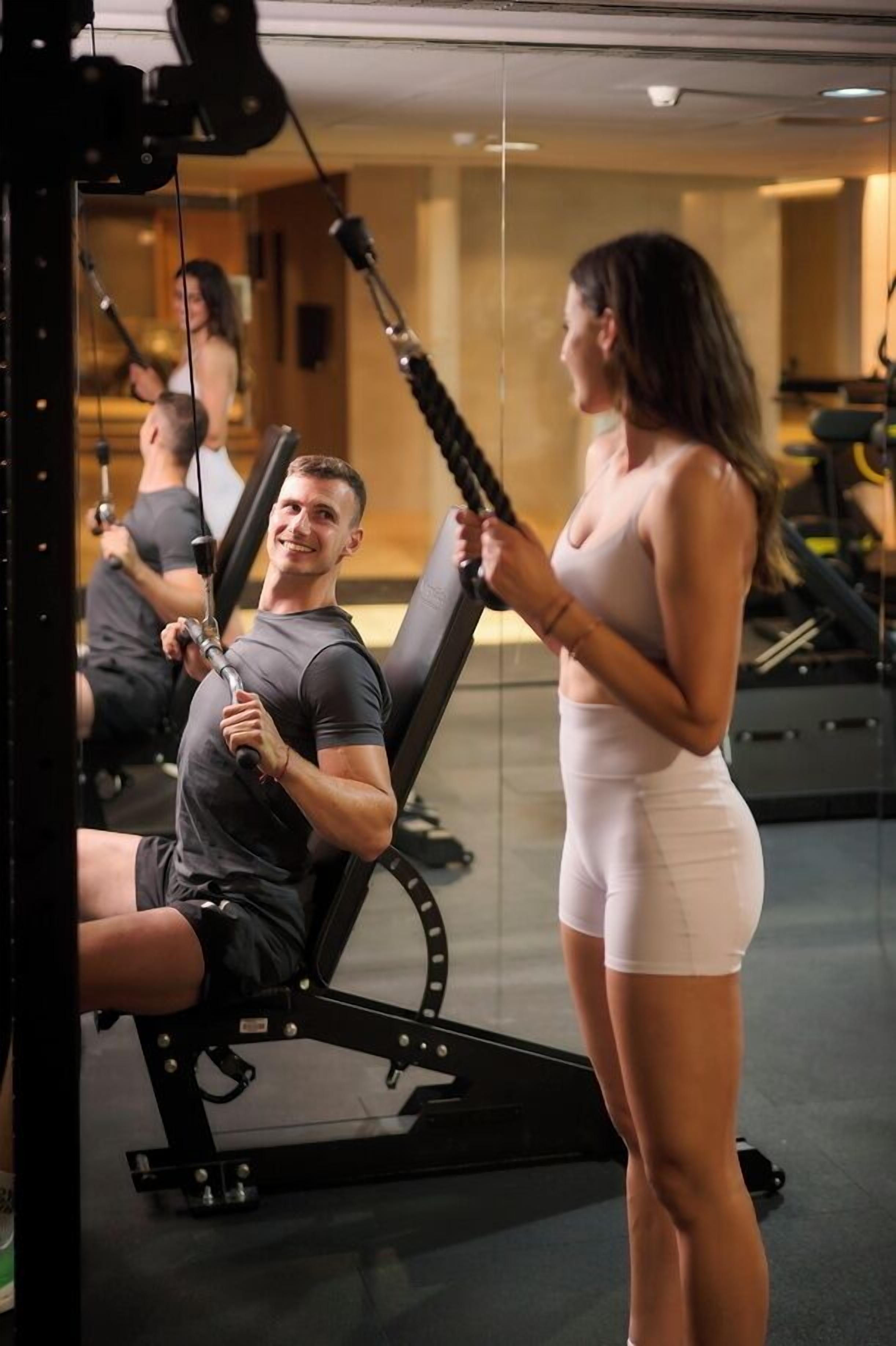 Une salle de sport équipée de matériel d’exercice, d’un miroir et où une personne utilise un appareil à câbles.