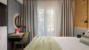 A modern hotel room with a large bed, a desk with a chair, a mirror, and a view of the outdoors through sheer curtains.