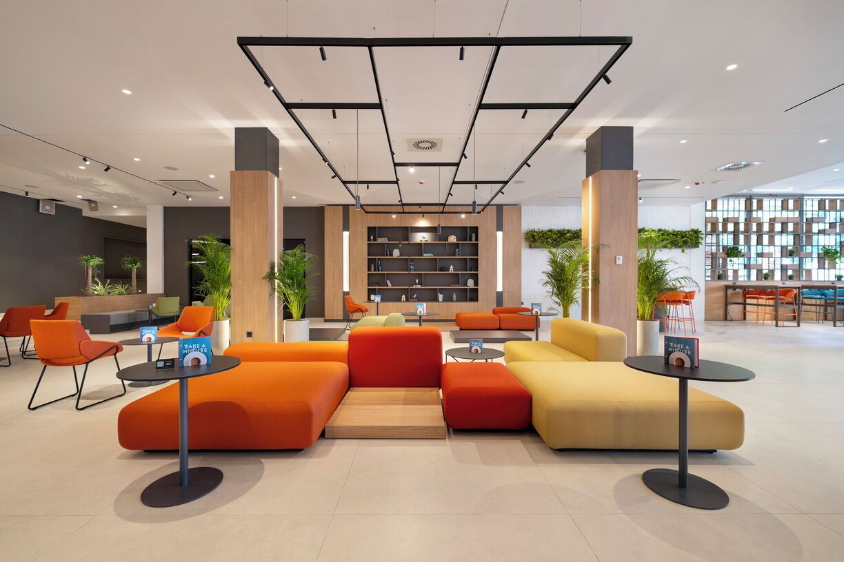 A modern lobby with orange and yellow seating, a central bookshelf, and a large window.
