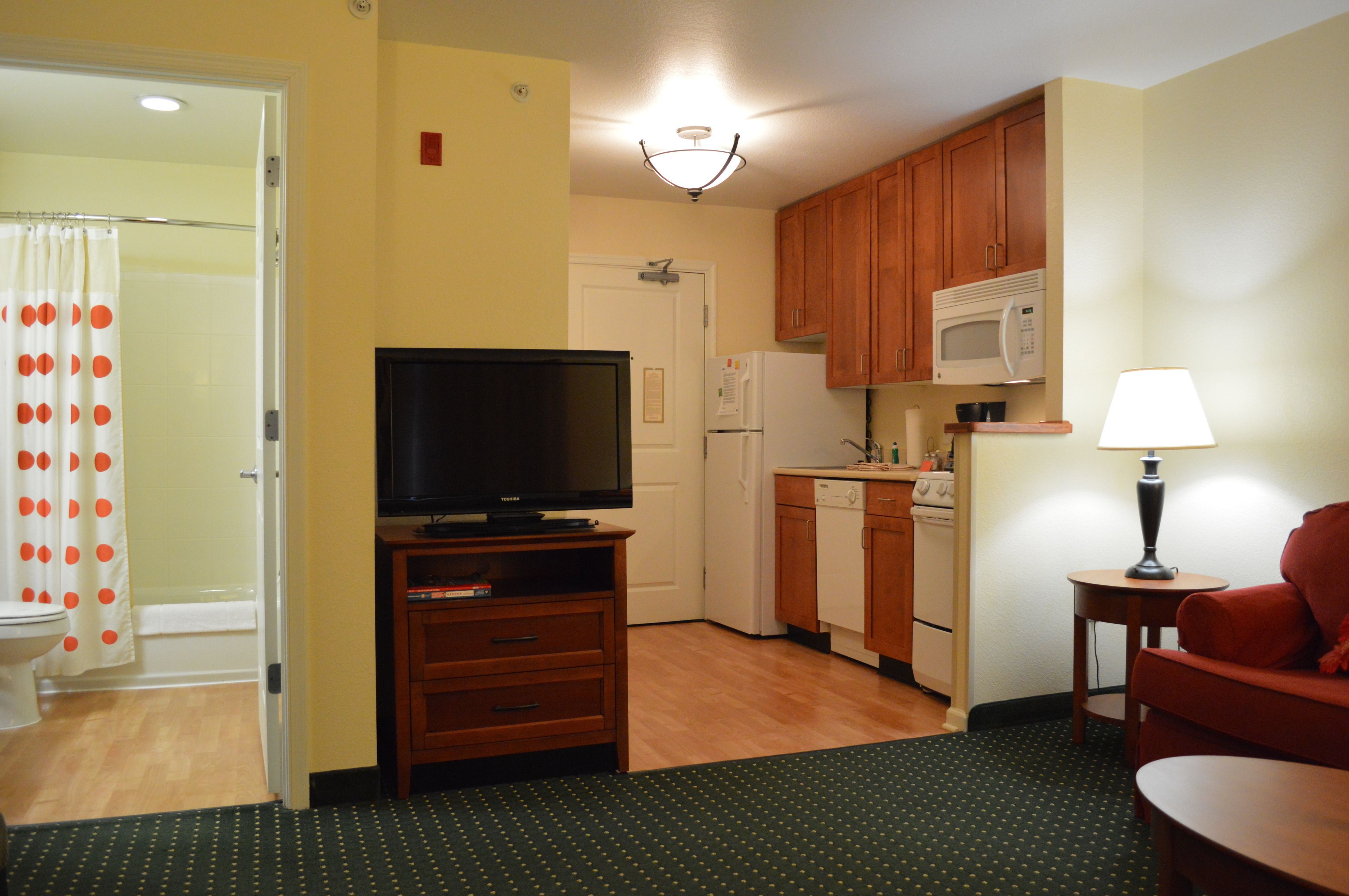 A compact kitchen with wooden cabinets, a microwave, a refrigerator, and a television on a wooden stand.