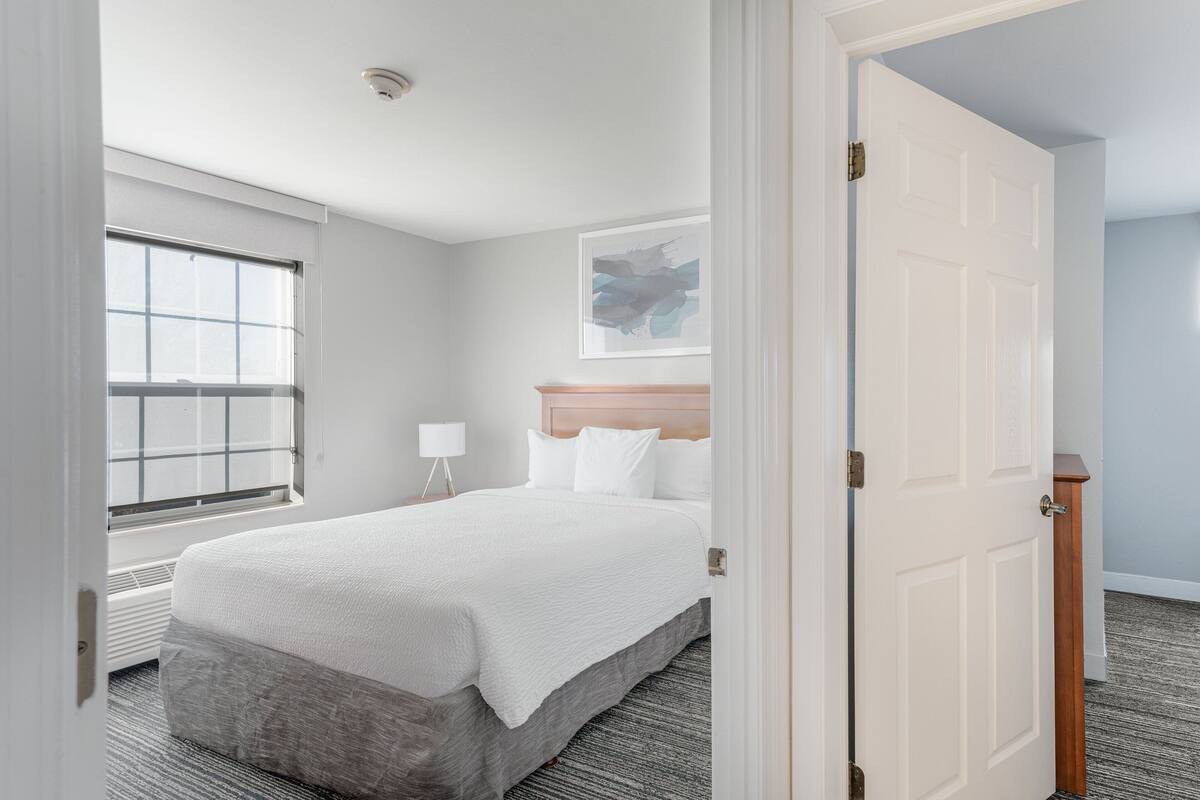 Une chambre d’hôtel avec un grand lit, une fenêtre, une table de chevet avec une lampe et un tableau encadré au mur.