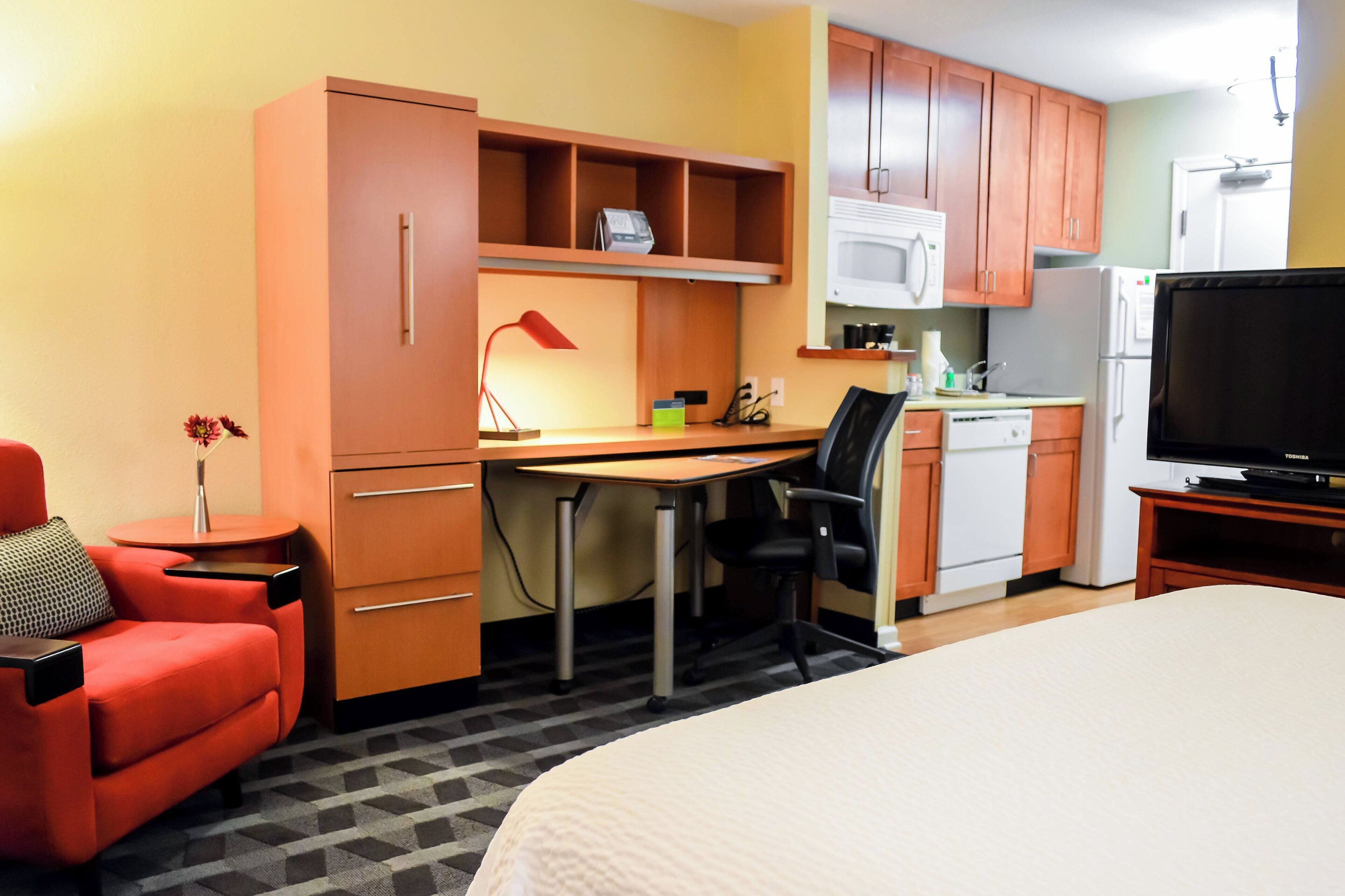 A compact hotel room with a bed, a desk, a chair, a TV, a kitchenette, and a small seating area with a chair and a table.