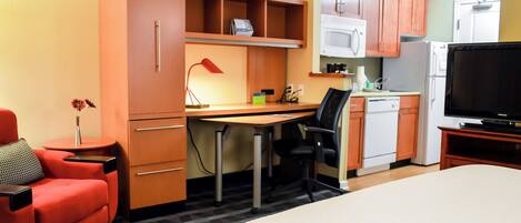 A compact hotel room with a bed, a desk, a chair, a TV, a kitchenette, and a small seating area with a chair and a table.