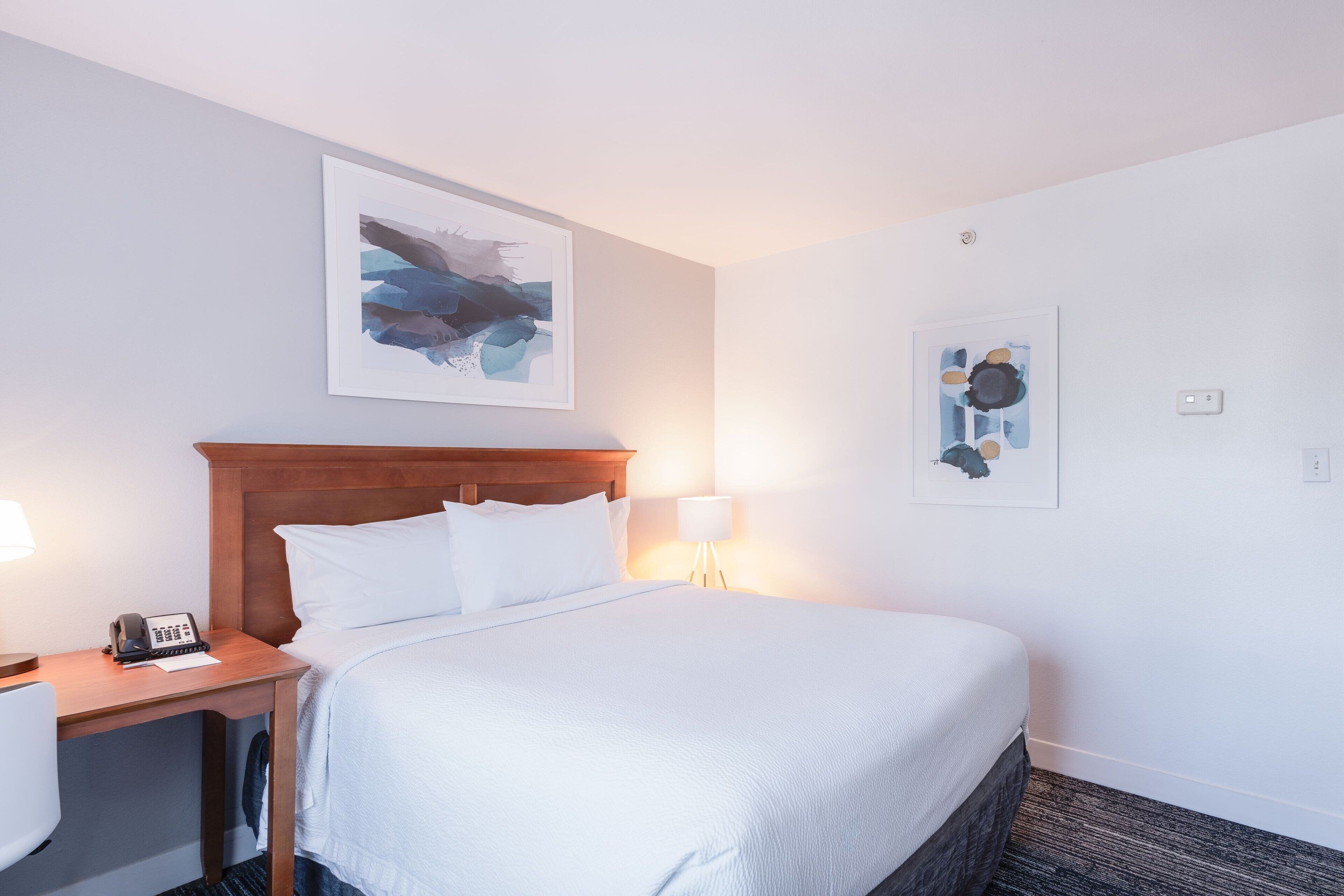 A hotel room with a large bed, a wooden nightstand with a phone, a desk, and two wall-mounted lamps.