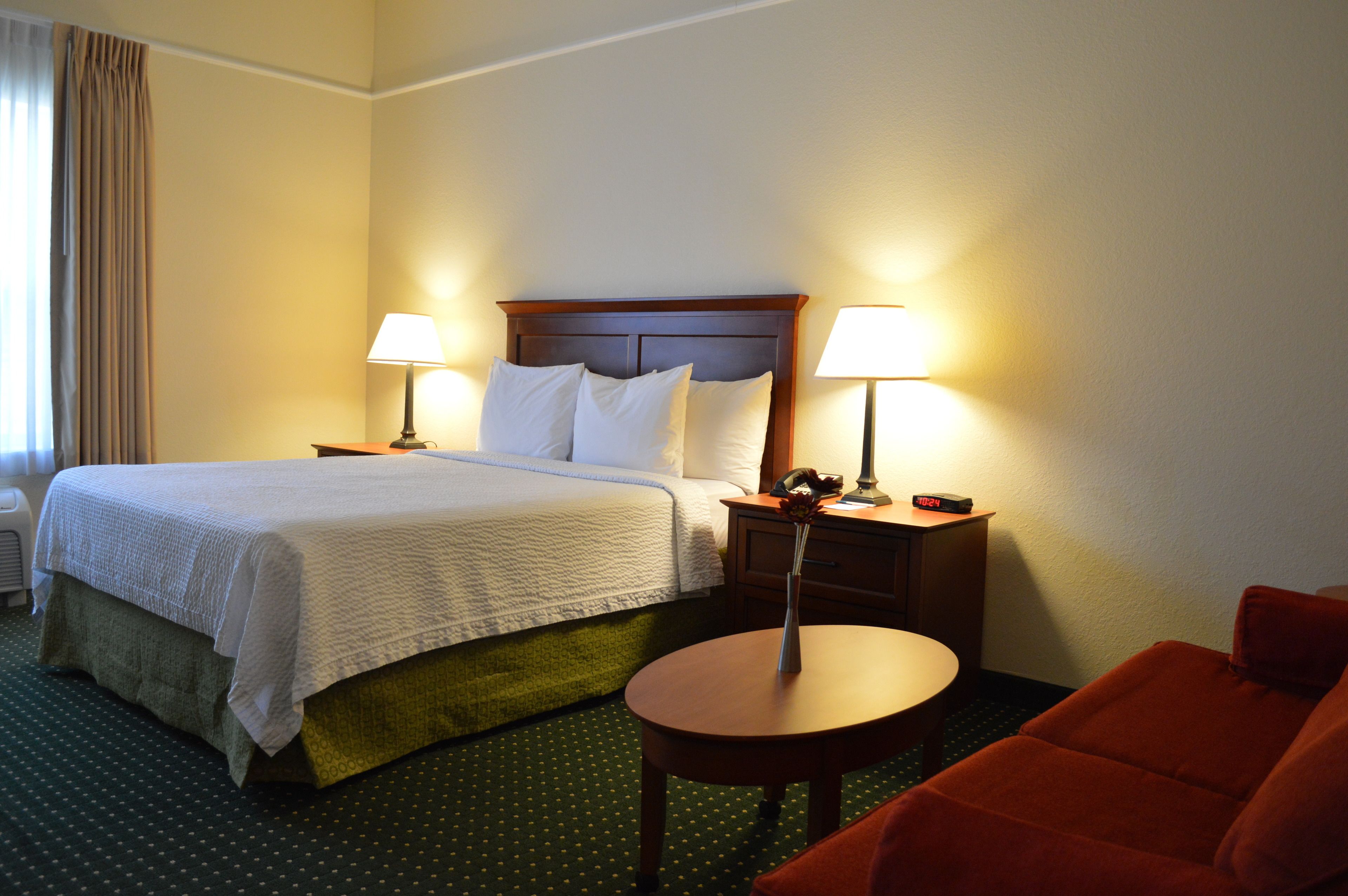 A hotel room with a bed, two bedside lamps, a nightstand with a phone and alarm clock, a small table, and a red armchair.