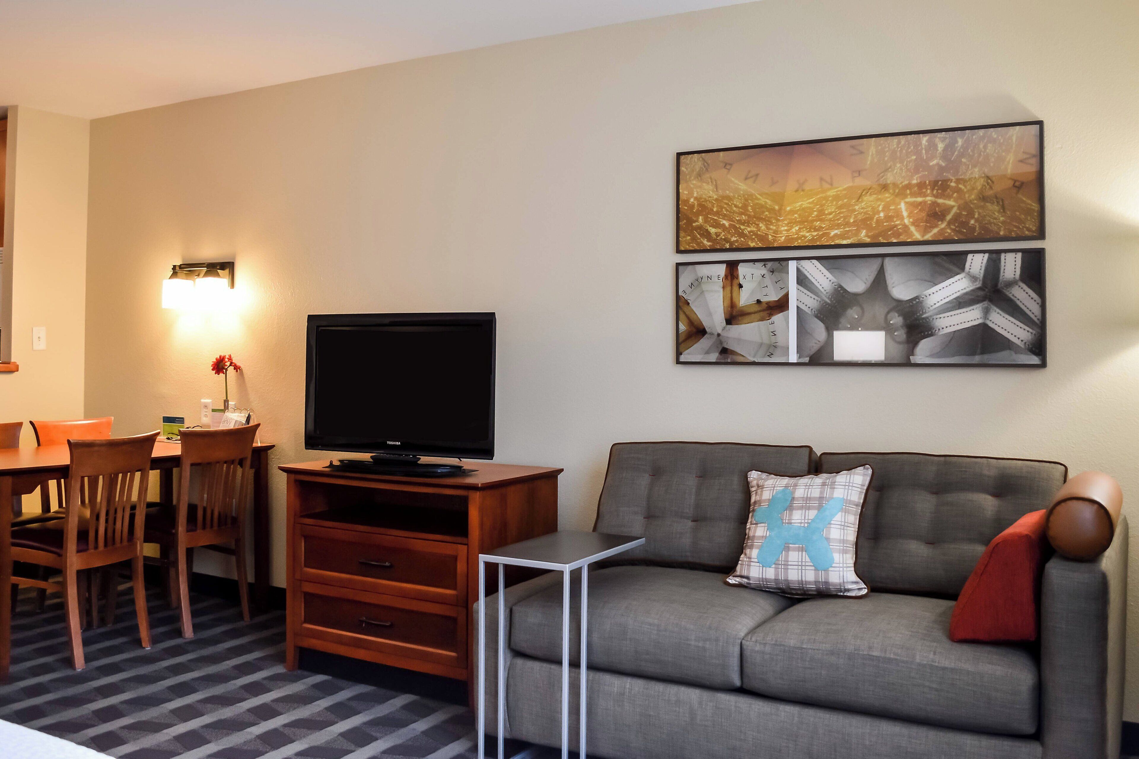 A living room with a flat-screen TV, a gray sofa with a blue pillow, a dining table with chairs, and wall art.