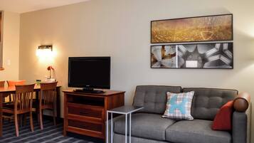 A living room with a flat-screen TV, a gray sofa with a blue pillow, a dining table with chairs, and wall art.