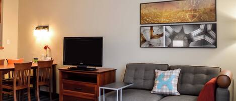 A living room with a flat-screen TV, a gray sofa with a blue pillow, a dining table with chairs, and wall art.