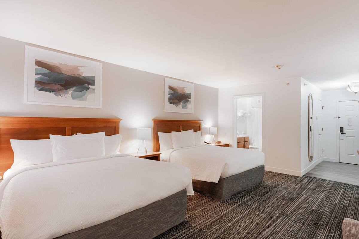 Une chambre d’hôtel avec deux lits, une tête de lit en bois, une moquette sombre et des œuvres d’art accrochées aux murs.