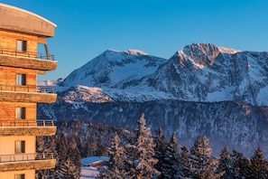 A multi-story building with balconies, set against a backdrop of snow-covered mountains and trees. - Noemys L'Ecrin des Neiges (Chamrousse)