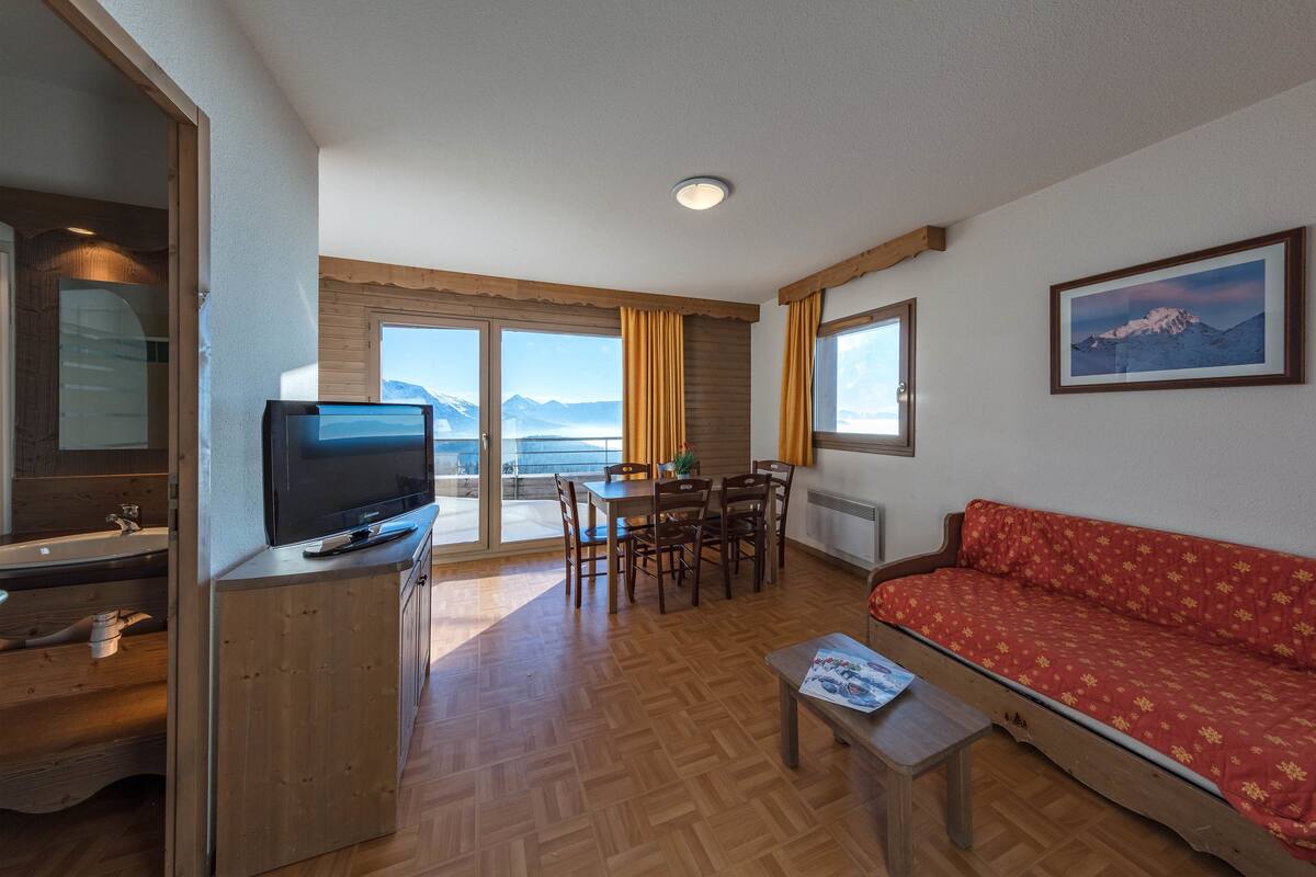 Una habitación con piso de madera, un sofá, un televisor, una mesa de comedor con sillas y un amplio ventanal con vista a la montaña.